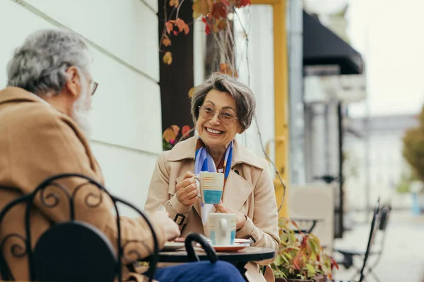 Cheerful senior woman in eyeglasses and trench coat holding cup during breakfast with husband on terrace of cafe — Stock Photo
