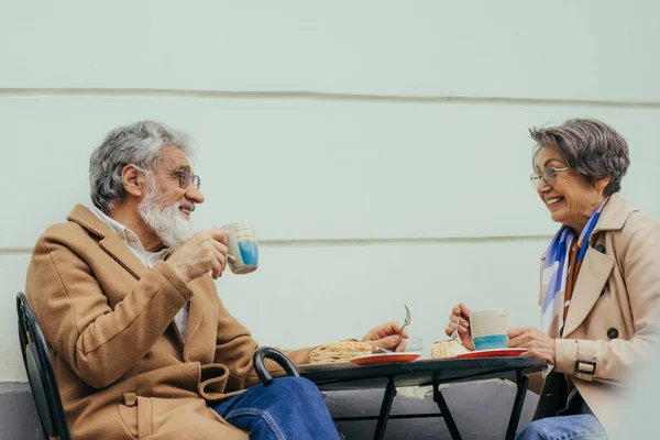 Donna più anziana allegra in occhiali da vista e trench che fa brunch con marito felice su terrazza di caffè — Foto stock