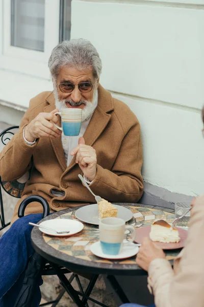 Uomo anziano barbuto in cappotto e occhiali bere tè vicino a torte e moglie sulla terrazza del caffè — Foto stock