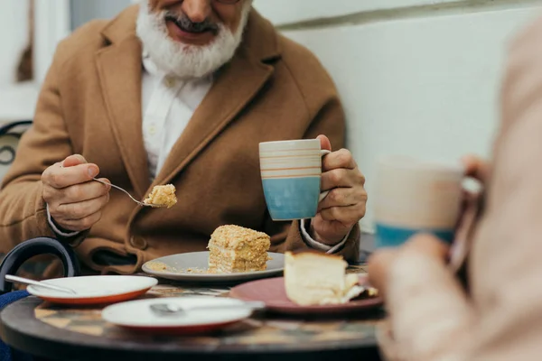 Vista recortada de hombre mayor alegre en abrigo sosteniendo taza de té y comer pastel cerca de la esposa en la terraza de la cafetería - foto de stock
