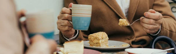 Vista recortada de hombre mayor en abrigo sosteniendo taza de té y comer pastel cerca de la esposa en la terraza de la cafetería, pancarta - foto de stock