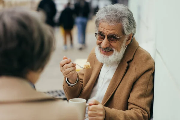 Happy senior man in coat holding cup of tea and eating cake near wife on terrace of cafe — Stock Photo