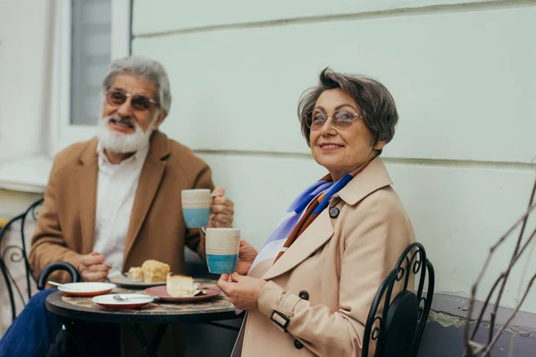 Feliz anciana en gafas de vista sosteniendo la taza mientras almorza con el marido barbudo en la terraza - foto de stock