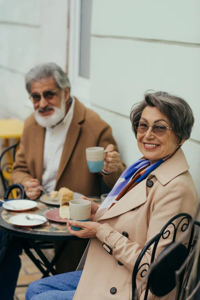 Happy senior woman in eyeglasses holding cup while having brunch with bearded husband on terrace of cafe — Stock Photo