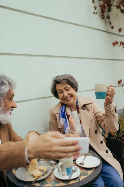 Bearded senior man taking selfie with cheerful wife during breakfast on terrace of cafe — Stock Photo