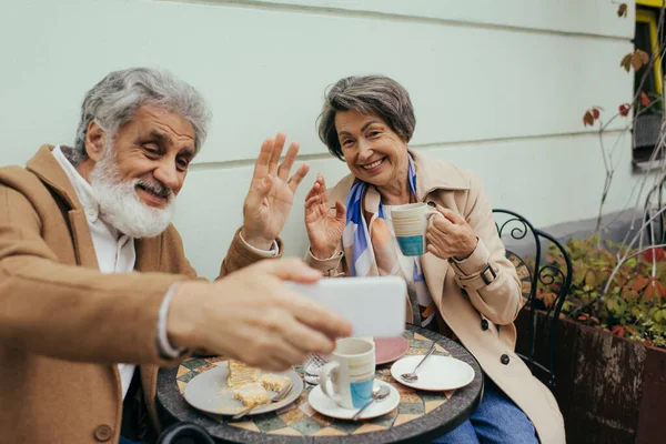 Coppia più anziana felice che ha la videochiamata e le mani ondulate durante colazione su terrazza di caffè — Foto stock