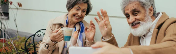 Feliz pareja de ancianos teniendo videollamadas y saludando las manos durante el desayuno en la terraza de la cafetería, pancarta - foto de stock