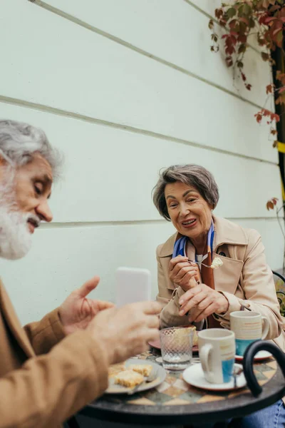 Bearded senior man showing smartphone to cheerful wife during breakfast on terrace of cafe — Stock Photo