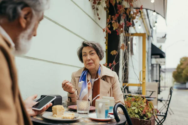 Glückliche Seniorin isst Kuchen und schaut Ehemann mit Smartphone auf Caféterrasse an — Stockfoto