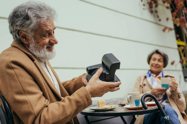 Hombre barbudo feliz sosteniendo la cámara vintage y tomando la foto de la mujer mayor borrosa durante el desayuno - foto de stock