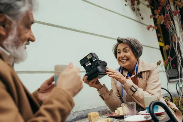 Счастливая пожилая женщина держит винтажную камеру возле размытого мужа с чашкой чая — стоковое фото