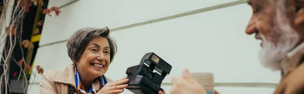 Heureuse femme âgée tenant caméra vintage près de mari flou avec tasse de thé, bannière — Photo de stock