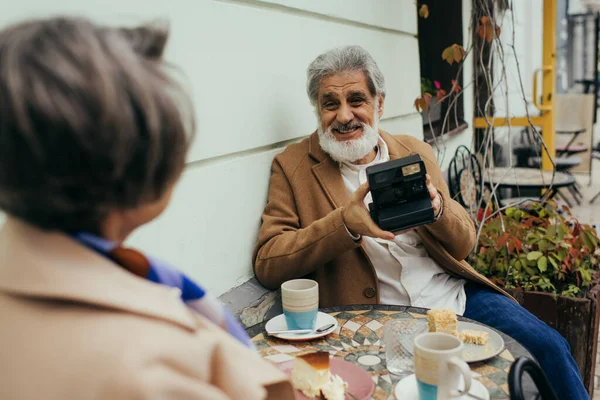 Щасливий старший чоловік з бородою тримає старовинну камеру під час сніданку з дружиною — стокове фото