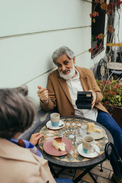 Vista ad alto angolo di felice uomo anziano con barba tenendo fotocamera vintage durante la colazione con moglie — Foto stock