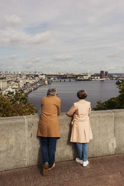Back view of retired couple in beige coats standing on bridge near river with city view — Stock Photo