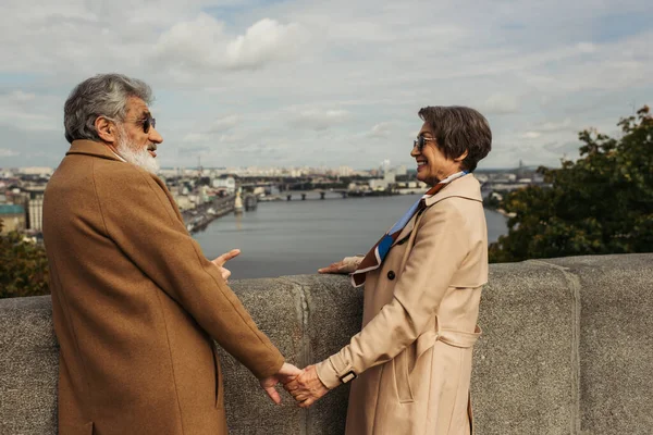 Pareja jubilada en abrigos beige tomados de la mano y mirándose el uno al otro cerca del río con vista a la ciudad - foto de stock