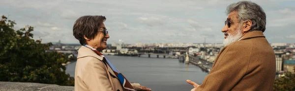 Retired couple in beige coats holding hands and looking at each other near river with city view, banner — Stock Photo