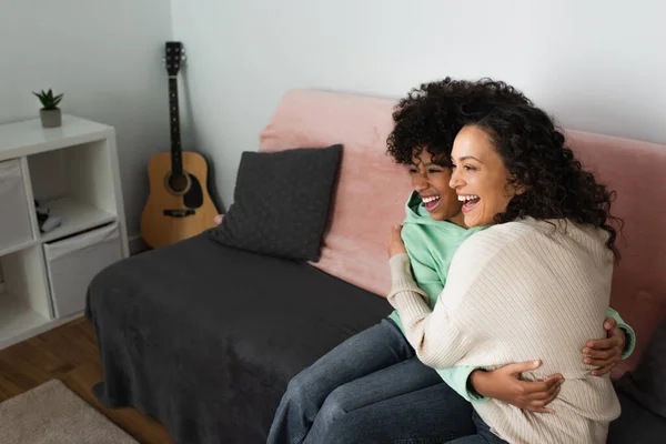 Positive african american mother and daughter hugging while sitting on couch — Stock Photo