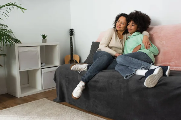 Feliz africano americano madre abrazando alegre preadolescente hija en sala de estar - foto de stock