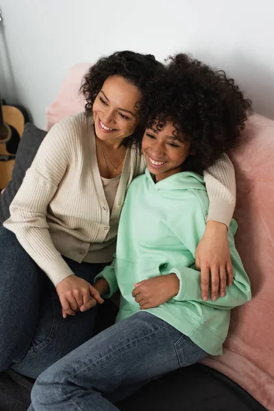 Vista de ángulo alto de la madre afroamericana feliz abrazando alegre hija preadolescente en la sala de estar - foto de stock