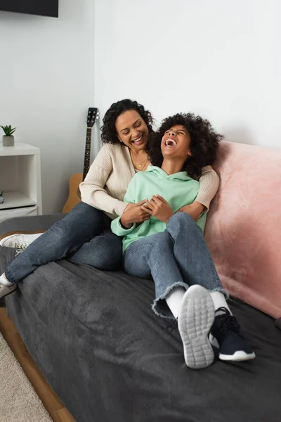 Happy african american mother tickling cheerful preteen daughter in living room — Stock Photo