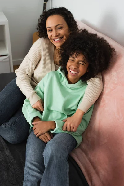 Vista ad alto angolo di felice madre afro-americana che abbraccia sorridente figlia preadolescente in soggiorno — Foto stock