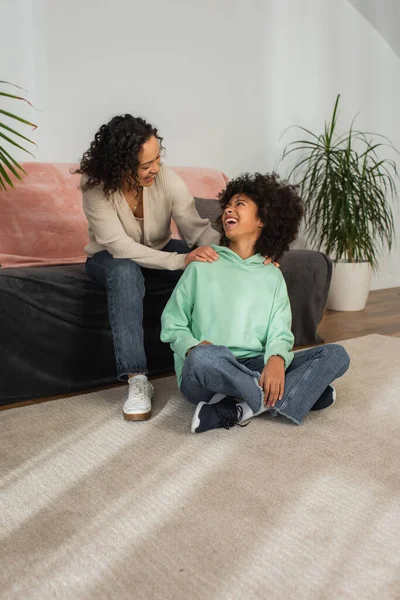 Encaracolado afro-americano mãe olhando animado pré-adolescente filha sentado com pernas cruzadas no tapete — Fotografia de Stock