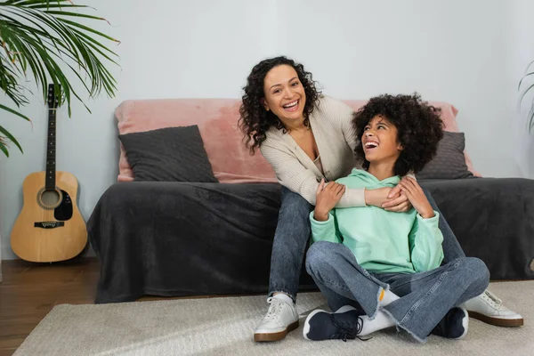 Lockige afrikanisch-amerikanische Mutter lächelt, während sie aufgeregte Tochter umarmt, die mit überkreuzten Beinen auf dem Teppich sitzt — Stockfoto