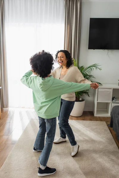 Excitada mujer afroamericana cogida de la mano con su hija preadolescente rizada mientras baila en casa - foto de stock