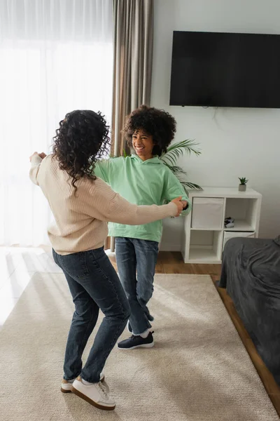Feliz africana americana chica cogida de la mano con rizado madre mientras bailando en casa - foto de stock
