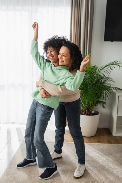 Piena lunghezza di allegra madre afro-americana che abbraccia la figlia preadolescente con le mani sollevate — Foto stock