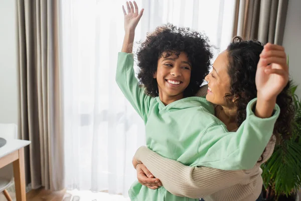 Joyeuse femme afro-américaine étreignant heureuse fille préadolescente avec les mains levées — Photo de stock