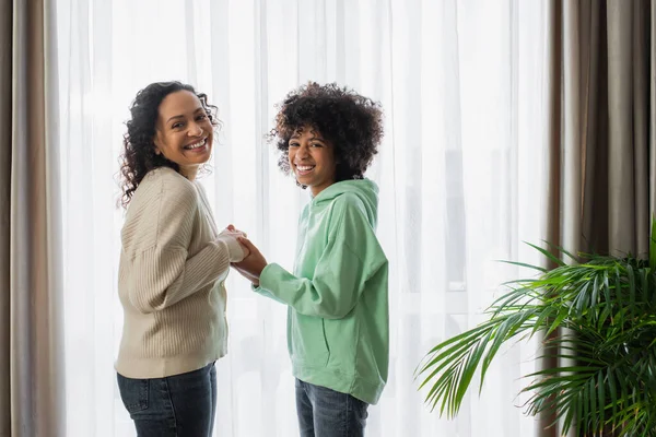 Curly african american woman smiling while holding hands with cheerful preteen daughter — Stock Photo