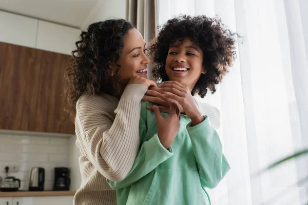 Baixo ângulo vista de mulher afro-americana satisfeita sorrindo enquanto abraçando alegre pré-adolescente filha — Fotografia de Stock