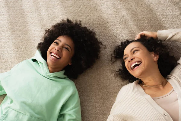 Vista superior de la chica afroamericana feliz sonriendo mientras está acostado en la alfombra con la madre alegre - foto de stock