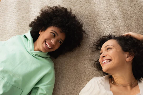 Vue du haut de heureuse fille afro-américaine souriant tout en regardant la mère gaie et couché sur le tapis — Photo de stock