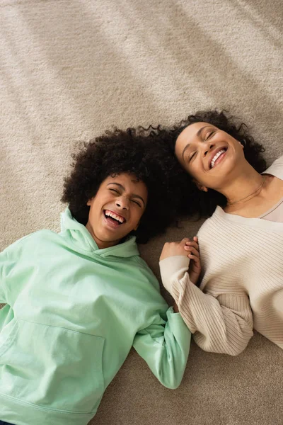 Vista superior de la chica afroamericana feliz sonriendo mientras está acostado en la alfombra con la madre positiva - foto de stock