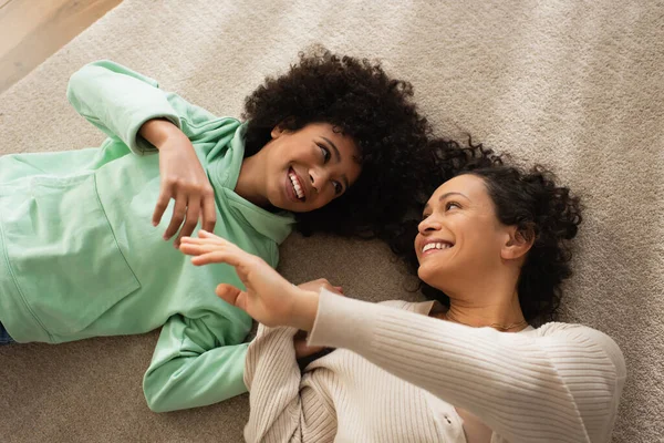 Vue du haut de joyeuse fille afro-américaine souriant tout en étant allongé sur le tapis avec une mère joyeuse — Photo de stock