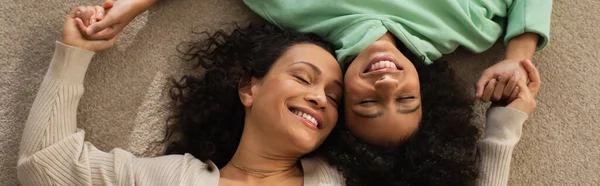 Top view of smiling african american girl lying on carpet with cheerful mother, banner — Stock Photo