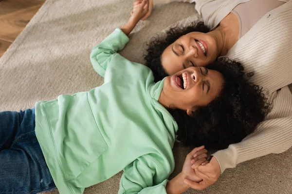 Ansicht von oben: fröhliches afrikanisch-amerikanisches Mädchen lacht, während es mit fröhlicher Mutter auf Teppich liegt — Stockfoto