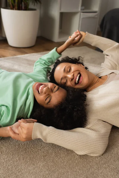 Menina americana africana feliz rindo enquanto deitado no tapete com mãe alegre — Fotografia de Stock