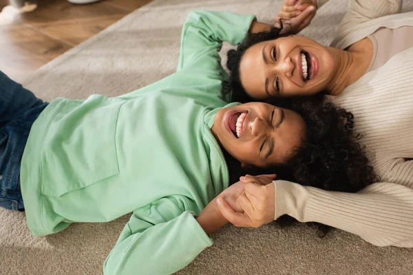 Positive Afroamerikanerin lacht, während sie mit fröhlicher Mutter auf Teppich liegt — Stockfoto