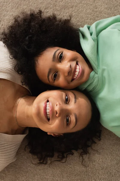 Blick von oben auf glückliches afrikanisch-amerikanisches Mädchen, das mit fröhlicher Mutter auf Teppich liegt und in die Kamera blickt — Stockfoto