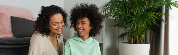 Happy african american preteen girl looking at curly mother in living room, banner — Stock Photo