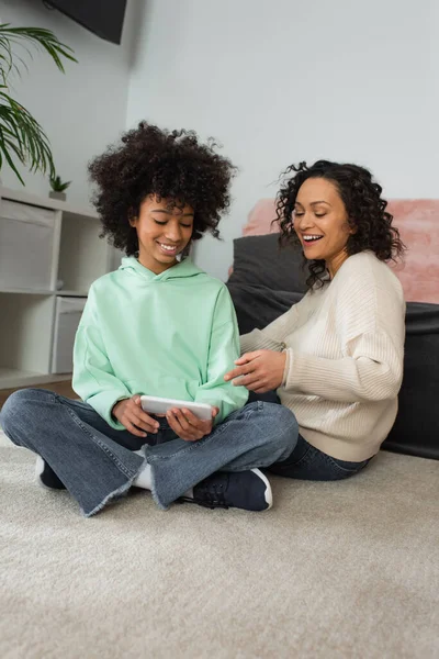 Веселая африканская американская мать, указывающая на смартфон в руках девочки-подростка, сидящей со скрещенными ногами на ковре — стоковое фото