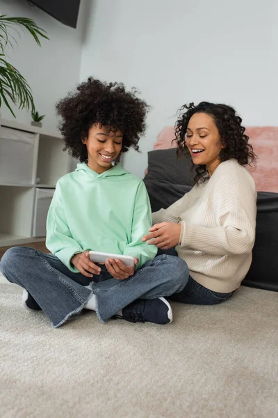 Fröhliche afrikanisch-amerikanische Mutter zeigt auf Smartphone in den Händen eines frühgeborenen Mädchens, das mit überkreuzten Beinen auf Teppich sitzt — Stockfoto