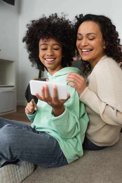 Feliz afroamericano preadolescente chica sosteniendo smartphone cerca de la madre positiva - foto de stock