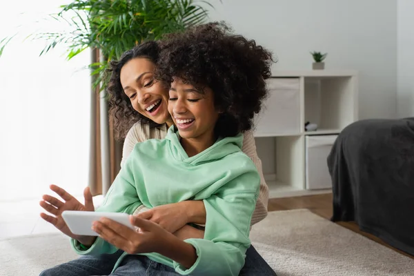 Menina pré-adolescente americana africana feliz e mãe encaracolada usando telefone celular na sala de estar — Fotografia de Stock