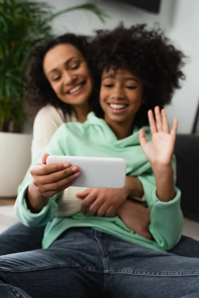 Feliz afroamericano preadolescente chica agitando la mano cerca de la madre rizada durante la videollamada en el teléfono inteligente - foto de stock