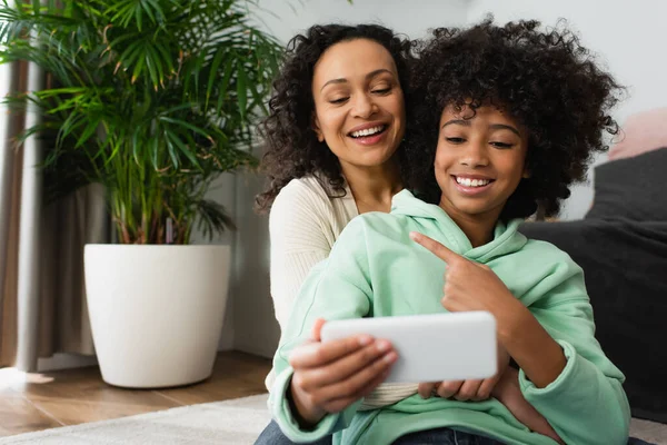 Happy african american preteen girl pointing at mother while taking selfie on smartphone — Stock Photo